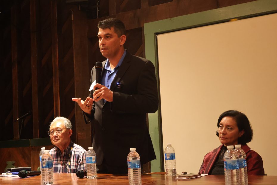 A man in a blue shirt and blazer speaks into a microphone at a panel discussion. Two other individuals, a man and a woman, sit on either side of him at the table with water bottles in front of them.