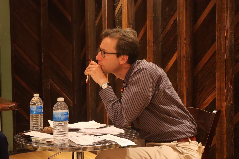 A man in glasses and a striped shirt sits at a table holding a pen to his mouth, with water bottles and papers in front of him. He appears to be deep in thought.