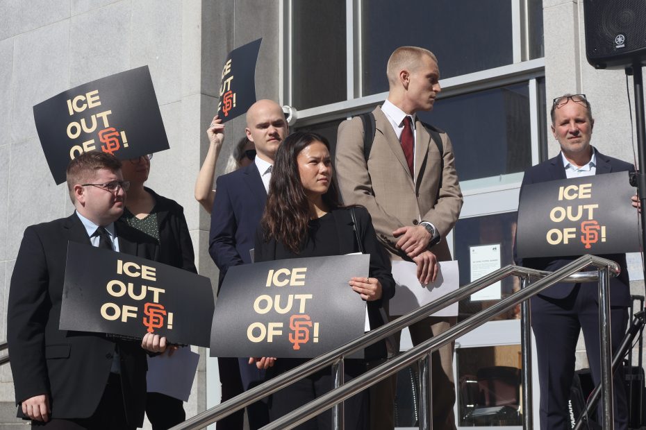 A group of people stands on building steps holding signs that read "ICE OUT OF SF!" in a protest or rally. Some participants are dressed in formal attire.