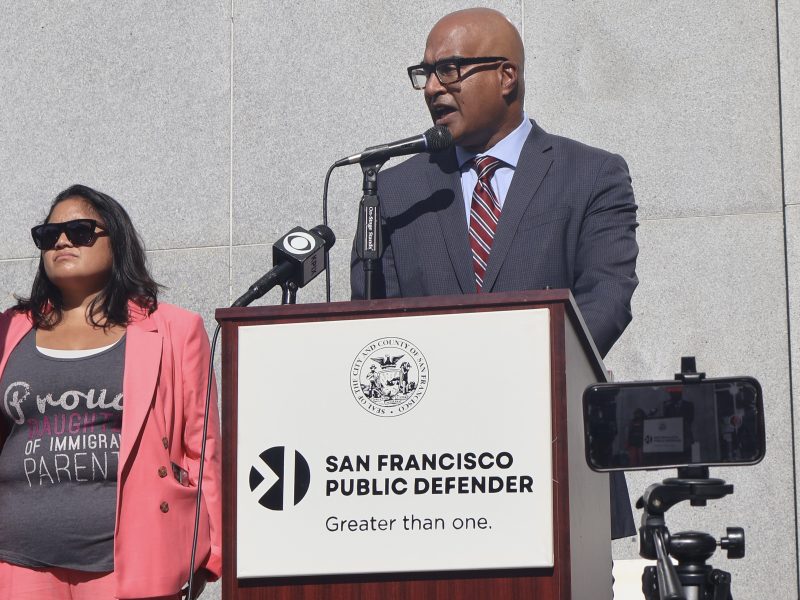 A person in a suit speaks at a podium labeled "San Francisco Public Defender." Another person stands nearby, wearing sunglasses and a shirt that reads "Proud daughter of immigrant parents.