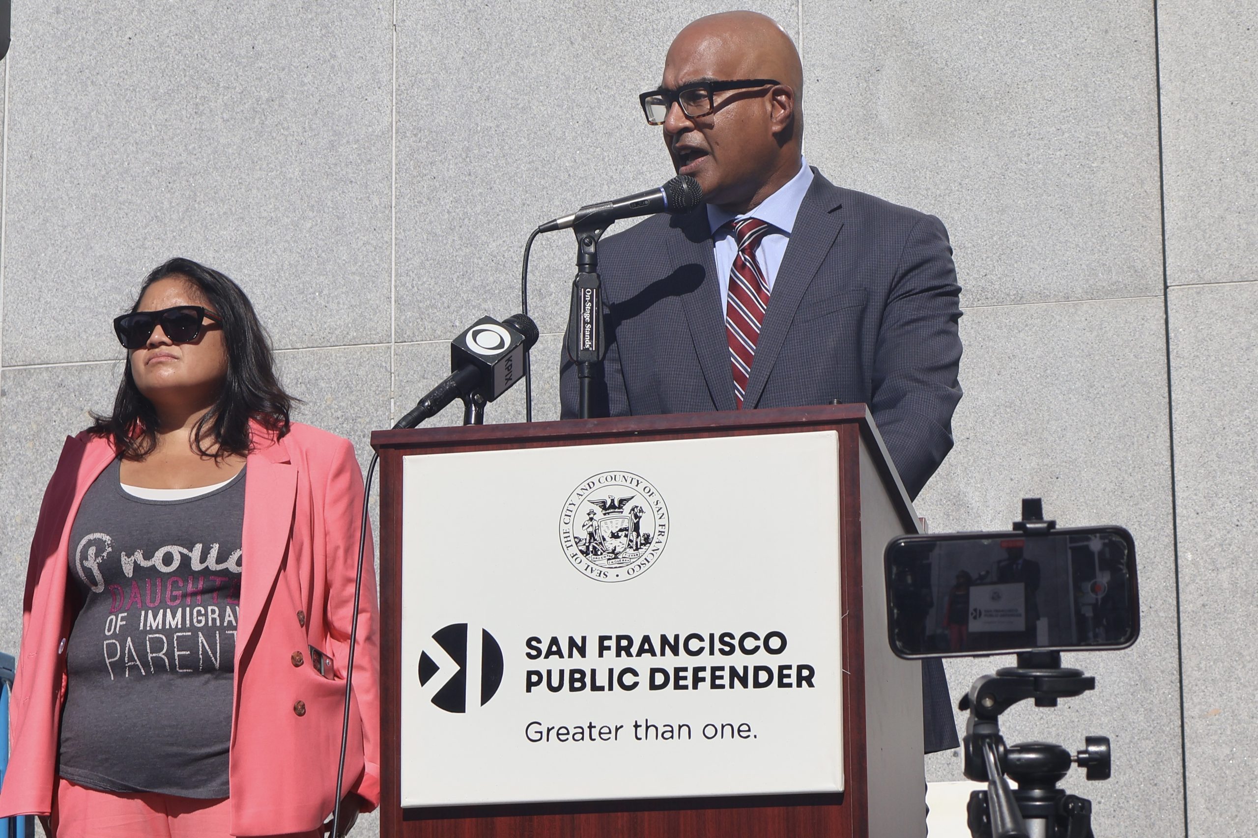 A person in a suit speaks at a podium labeled "San Francisco Public Defender." Another person stands nearby, wearing sunglasses and a shirt that reads "Proud daughter of immigrant parents.
