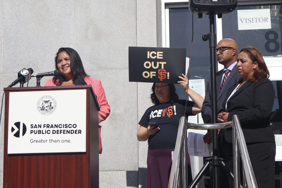 A woman speaks at a podium outside the San Francisco Public Defender's Office. Next to her are three individuals, one holding a sign that reads "ICE OUT OF SF".