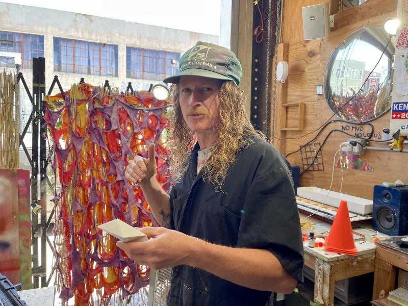 A person with long curly hair, wearing a cap and a black shirt, stands in a cluttered workspace holding a piece of paper, with colorful artwork and various objects in the background.