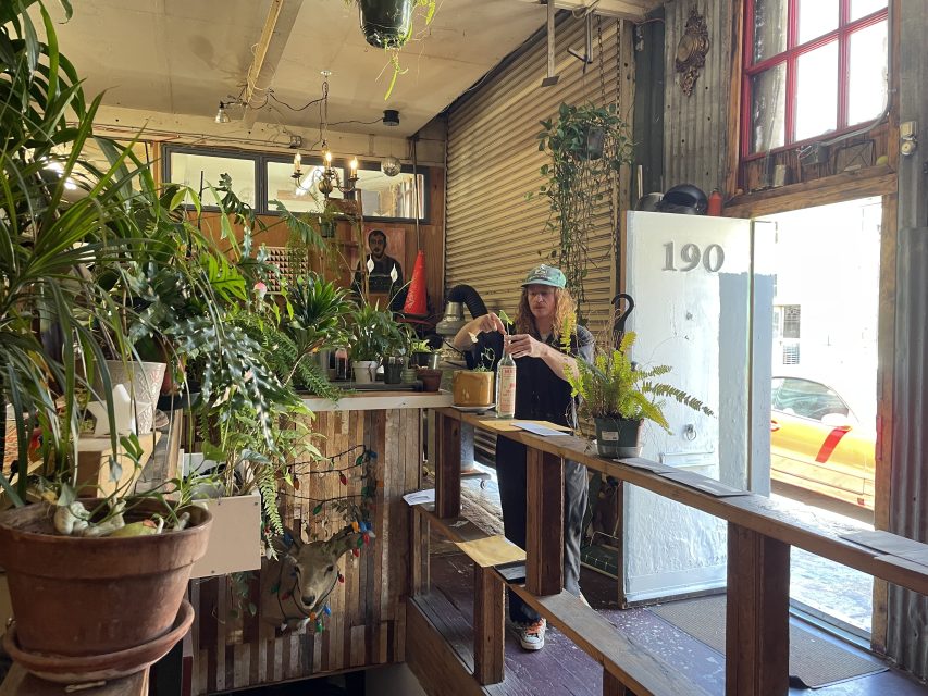 A person waters plants inside a rustic plant shop with wooden shelves filled with various potted plants. Another person stands behind the counter. The shop's entrance and street view are visible.