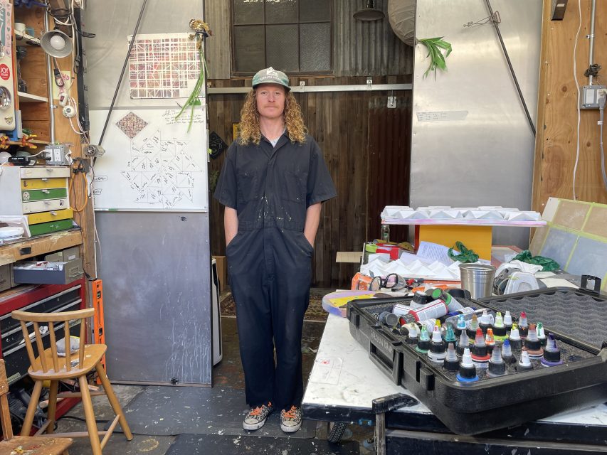 A person with long hair wearing a cap and dark coveralls stands in a cluttered workshop, with various tools and supplies on a table in the foreground.