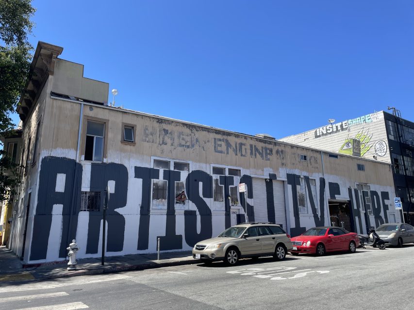 A building with a large mural reading "ARTISTS LIVE HERE" on its side. Three parked cars are visible in the foreground.