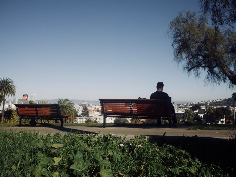 Two individuals sit on separate benches in a park, facing away from each other. The park features palm trees and greenery with a cityscape visible in the background under a clear sky.