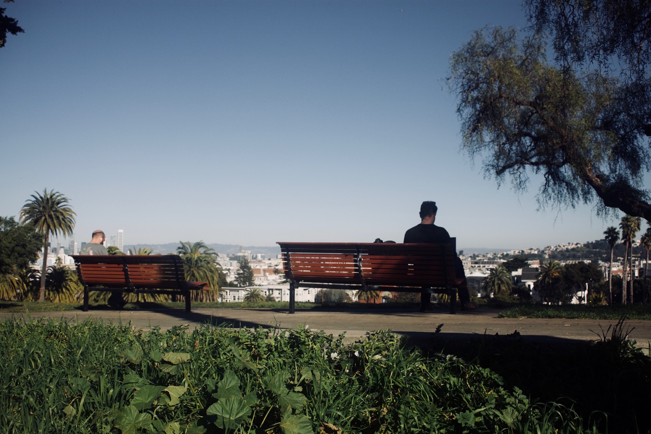 Two individuals sit on separate benches in a park, facing away from each other. The park features palm trees and greenery with a cityscape visible in the background under a clear sky.