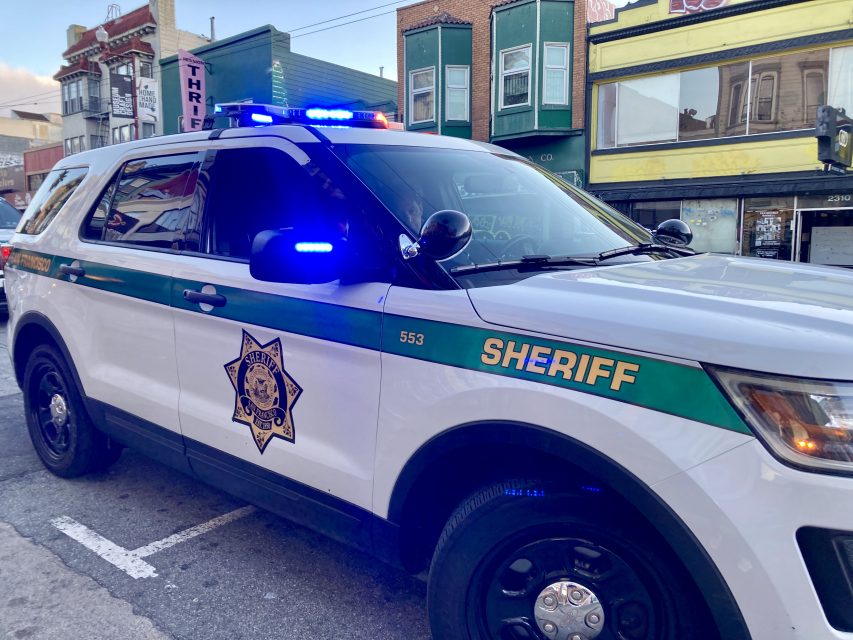A sheriff's SUV with flashing blue and red lights is parked on a street. The vehicle is white with green stripes and the word "SHERIFF" prominently displayed on its side.