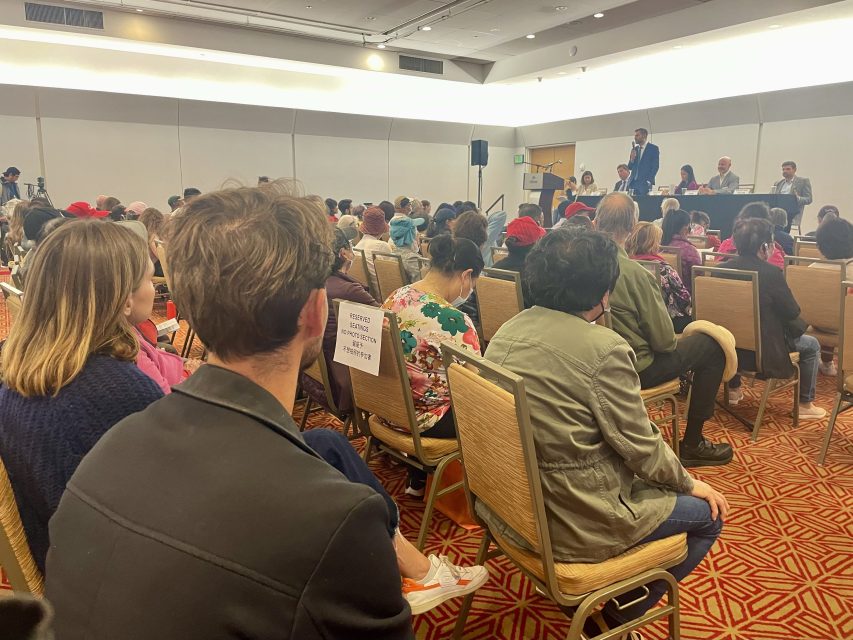 A crowded conference room with attendees seated, listening to a panel of speakers in the front. One attendee wears a mask and a sign reading "ATTENTION: COVID Prevention: Wear mask if unvaccinated.