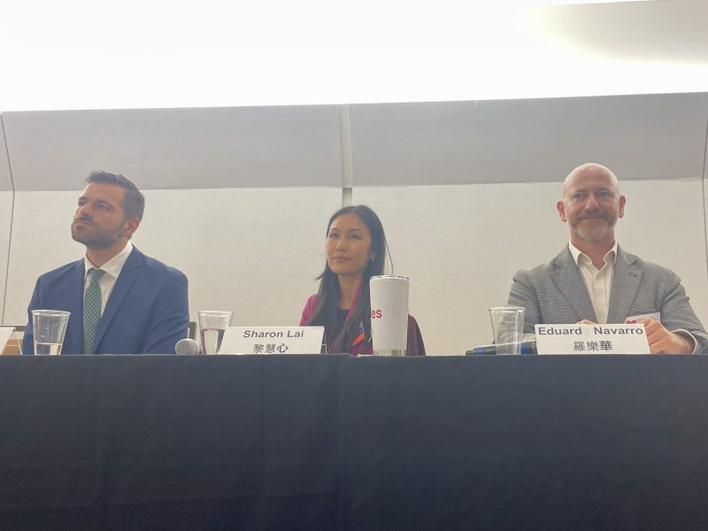 Three individuals are seated in front of a panel table with nameplates. The person on the left is wearing a suit and tie, the person in the middle has long hair, and the person on the right is bald and smiling.