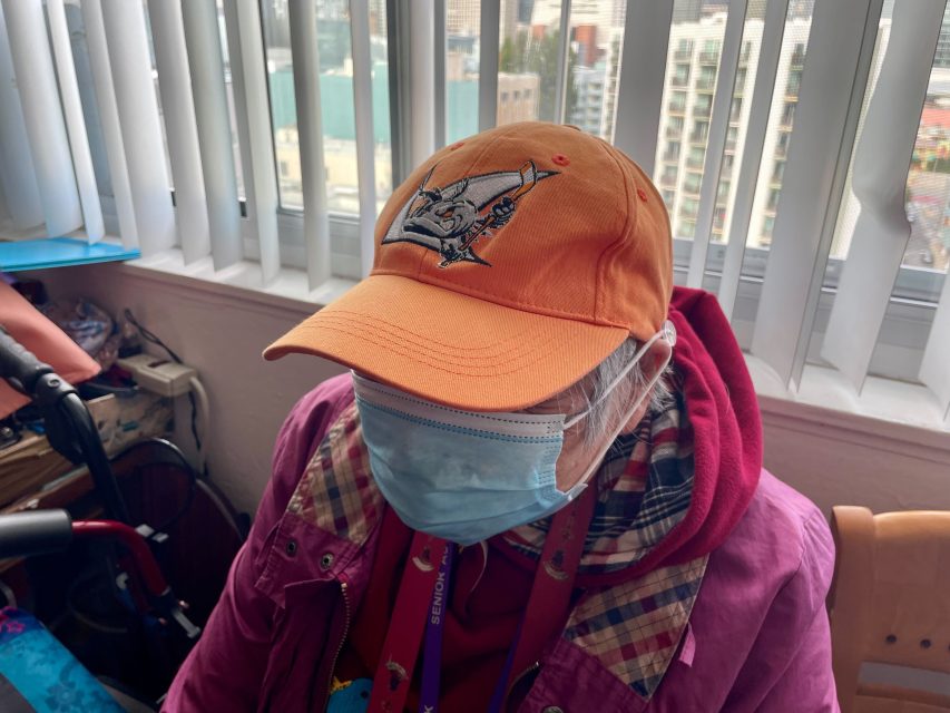 一個人戴著帶標誌的橙色帽子，藍色口罩，眼鏡，穿著粉色夾克，坐在一扇有豎百葉窗的窗戶前。