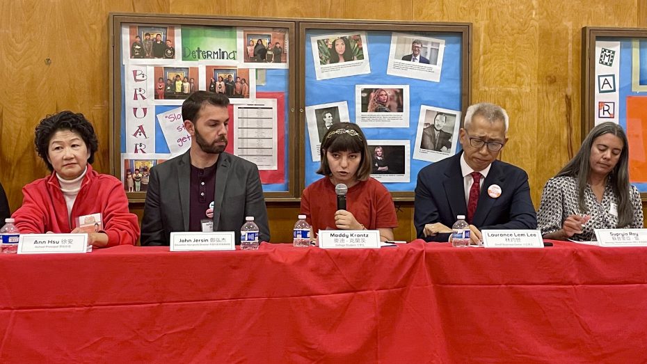 A panel of five people are seated at a table with red cloth, each with a microphone and name tag. The wall behind them displays various photos and documents. One individual in the center is speaking.
