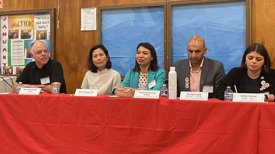 Five panelists sit at a table with a red tablecloth. Name plates in front of each person display their names. A blue backdrop is visible behind them and a bulletin board is partially seen on the left.