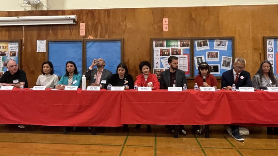A group of people sitting at a long table with a red tablecloth set in a hall, presumably at a formal meeting or event.