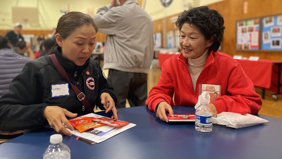 Two women sit at a table with documents in a community center. One woman points to a paper while the other looks on, both engaged in conversation.