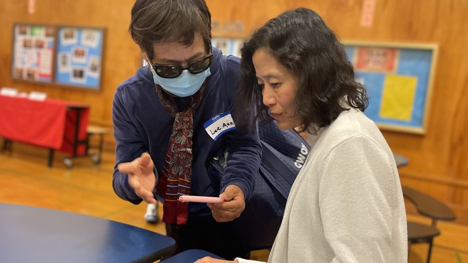 A person wearing sunglasses and a face mask shows something on a smartphone to another person in a white jacket. They are indoors, with bulletin boards and tables in the background.