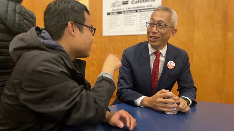 Two men are seated at a table engaging in conversation. One is holding a cup and appears to be listening intently. There is a sign on the wooden wall in the background.