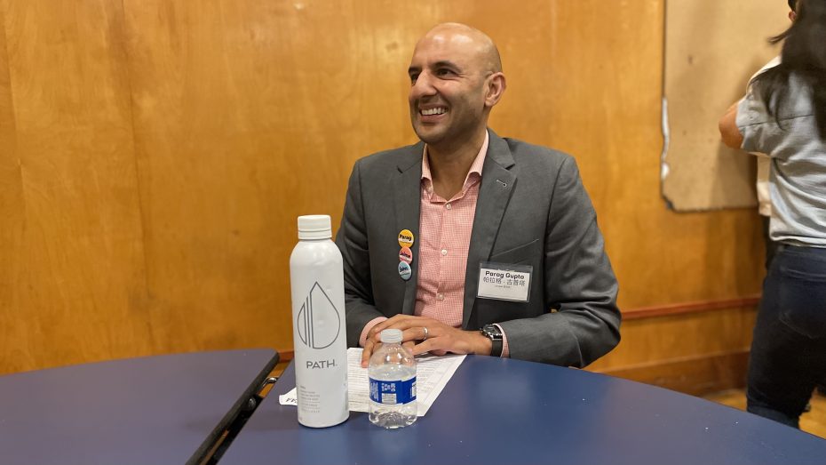 A man in a suit and pink shirt sits at a table with a Path water bottle and a small water bottle in front of him, smiling. He is wearing a name tag and has buttons pinned to his suit jacket.