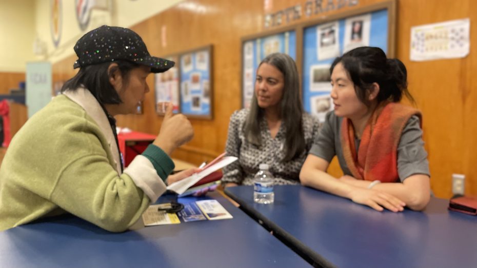 Three women engaged in a discussion while seated at a table in a brightly lit room. One woman is holding a notepad; another has a water bottle in front of her. Bulletin boards are visible in the background.