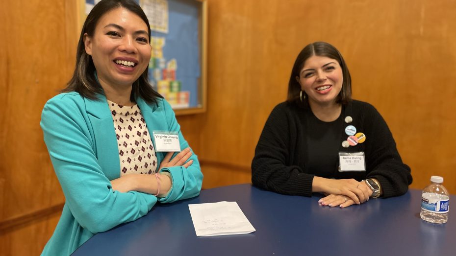 Two smiling women sitting at a blue table with a notepad, a water bottle, and a colorful background on the wall. Both women wear name tags and are engaged in conversation.