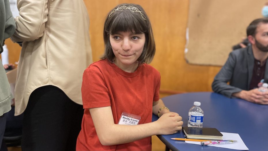 A person with short brown hair and a red shirt sits at a table with a notebook, pen, and water bottle in front of them.
