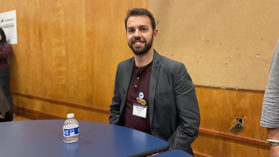 A man with a beard wearing a gray blazer and maroon shirt sits at a blue table with a bottle of water, smiling.