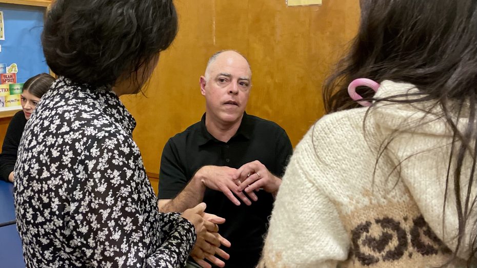 Three people in conversation, with one person gesturing while talking. Two have their backs to the camera, and one faces forward. They are in an indoor setting with a wooden backdrop.