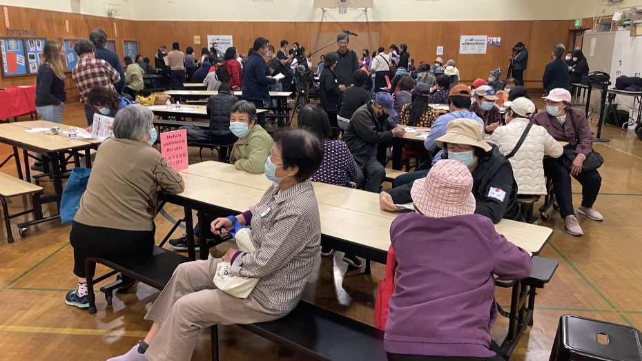 A large group of people, many wearing masks, are seated at tables and standing in line, gathered in a gymnasium for an event.