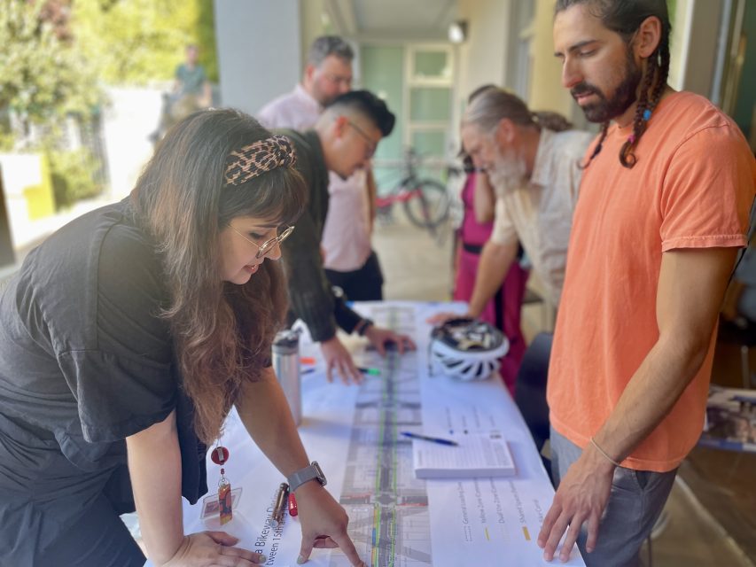 People engage in a discussion over a large map or document on a table. A woman points to a specific section while others observe or converse.