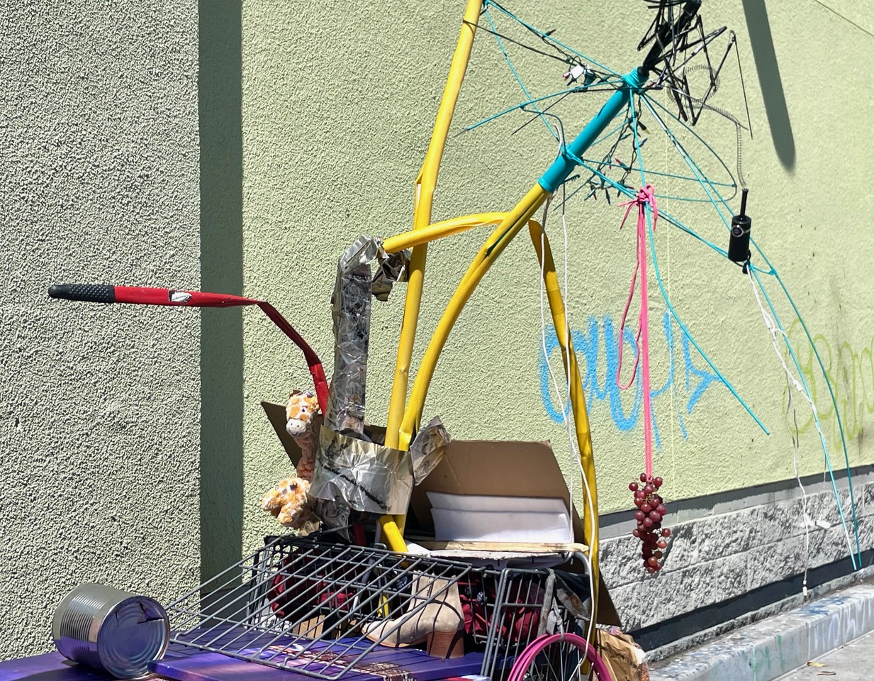A colorful abstract sculpture made from repurposed materials including pipes, wires, and a shopping cart is placed against a green wall. The sculpture is adorned with various decorations, including a bunch of fake grapes.