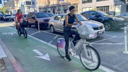 A woman rides a shared bike in a designated bike lane while looking at her phone. A cyclist follows behind her. Cars are parked and moving along the adjacent street.