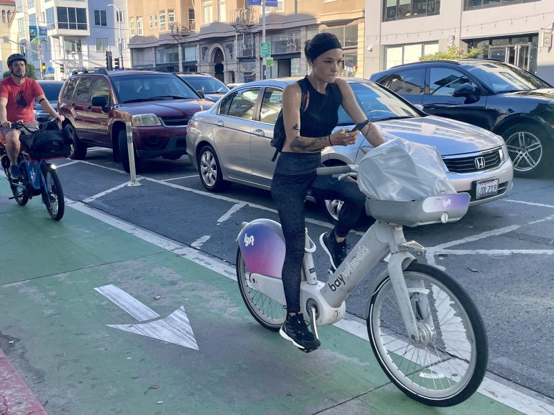 A woman rides a shared bike in a designated bike lane while looking at her phone. A cyclist follows behind her. Cars are parked and moving along the adjacent street.