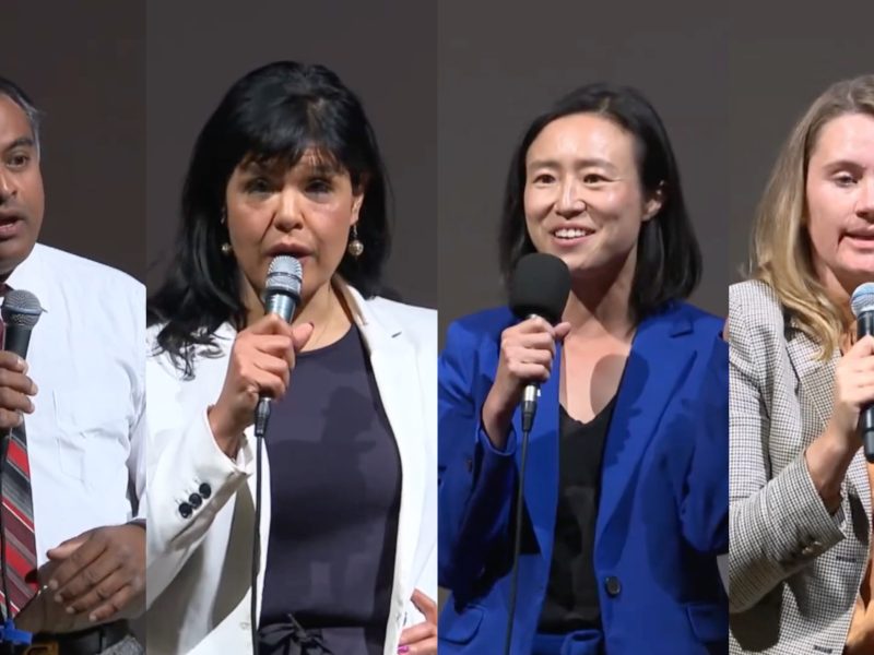 Four individuals stand side-by-side, each holding microphones and speaking. They are dressed in business attire and positioned against a plain backdrop.