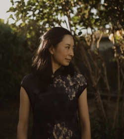 A person with shoulder-length dark hair stands outside in front of trees, wearing a dark floral-patterned dress, and looking to the left.