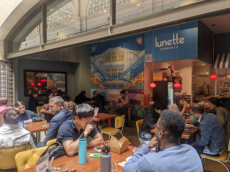 People dining at tables in a restaurant with a "lunette" sign and a mural on the wall in the background.