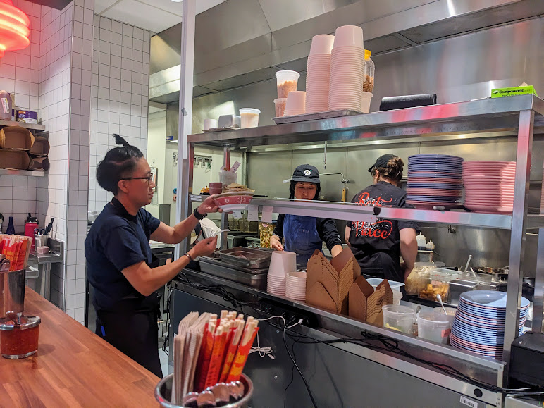 Three kitchen staff prepare food orders in a restaurant kitchen, organizing dishes and utensils. The workspace includes shelves stocked with containers, plates, and takeaway boxes.