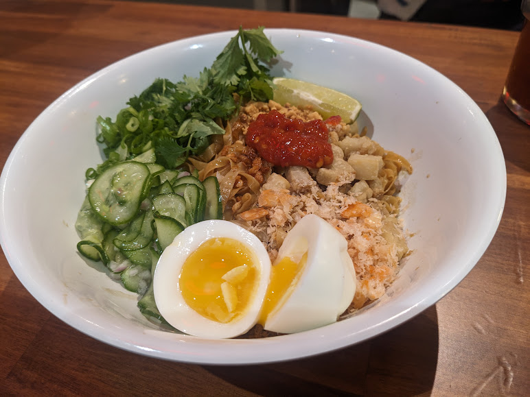 A white bowl containing noodles topped with sliced cucumbers, halved boiled eggs, leafy greens, a lime wedge, and a red paste, set on a wooden table.