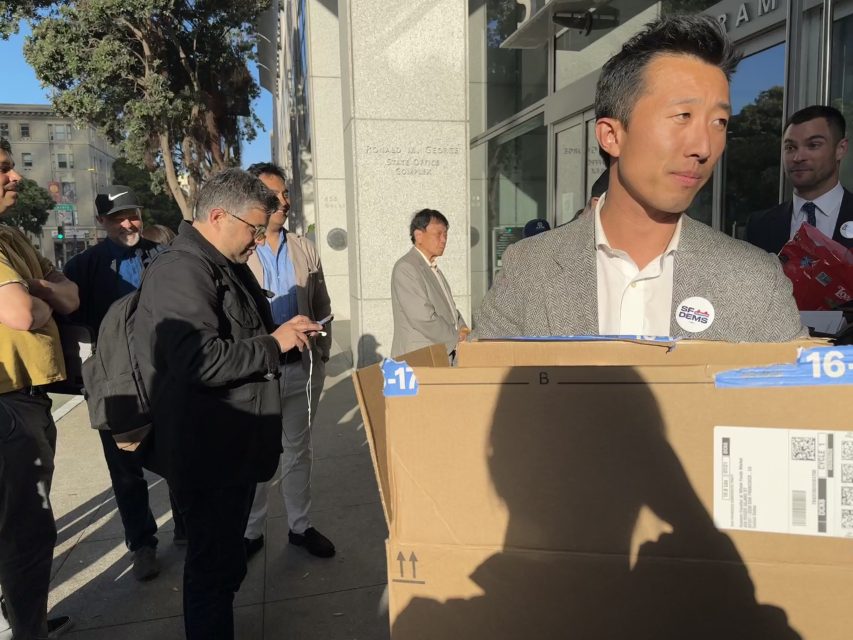 一名西裝男子在一棟玻璃門建築外抱著一個大紙箱。他周圍有幾個人站著，其中一人正在使用智能手機。