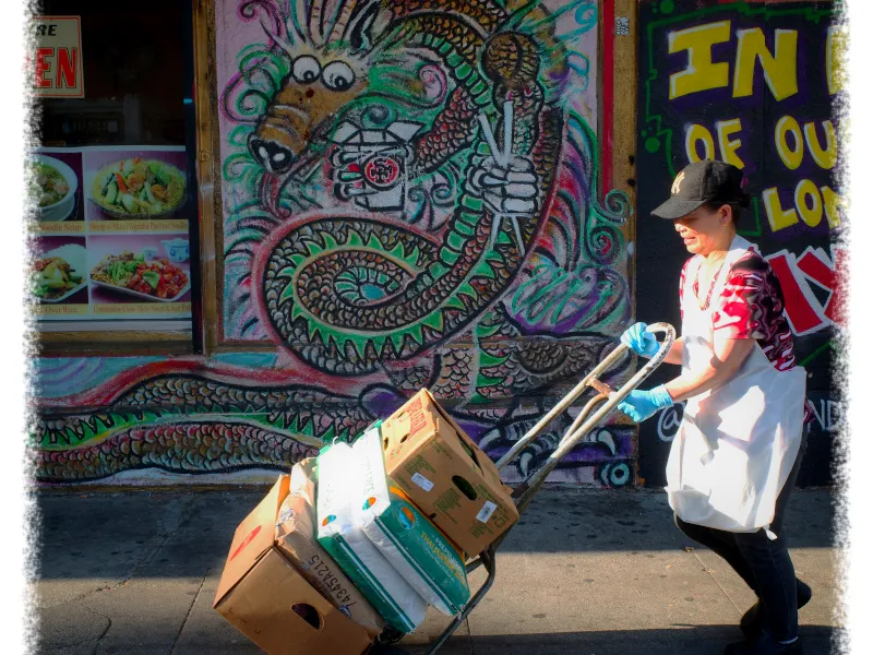 A person in a baseball cap and apron pushes a cart loaded with boxes past a colorful mural featuring a dragon on a street.