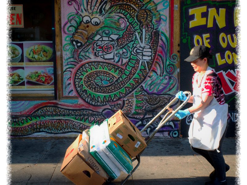 A person pushes a hand truck with boxes on a sidewalk in front of a storefront featuring a large dragon mural.