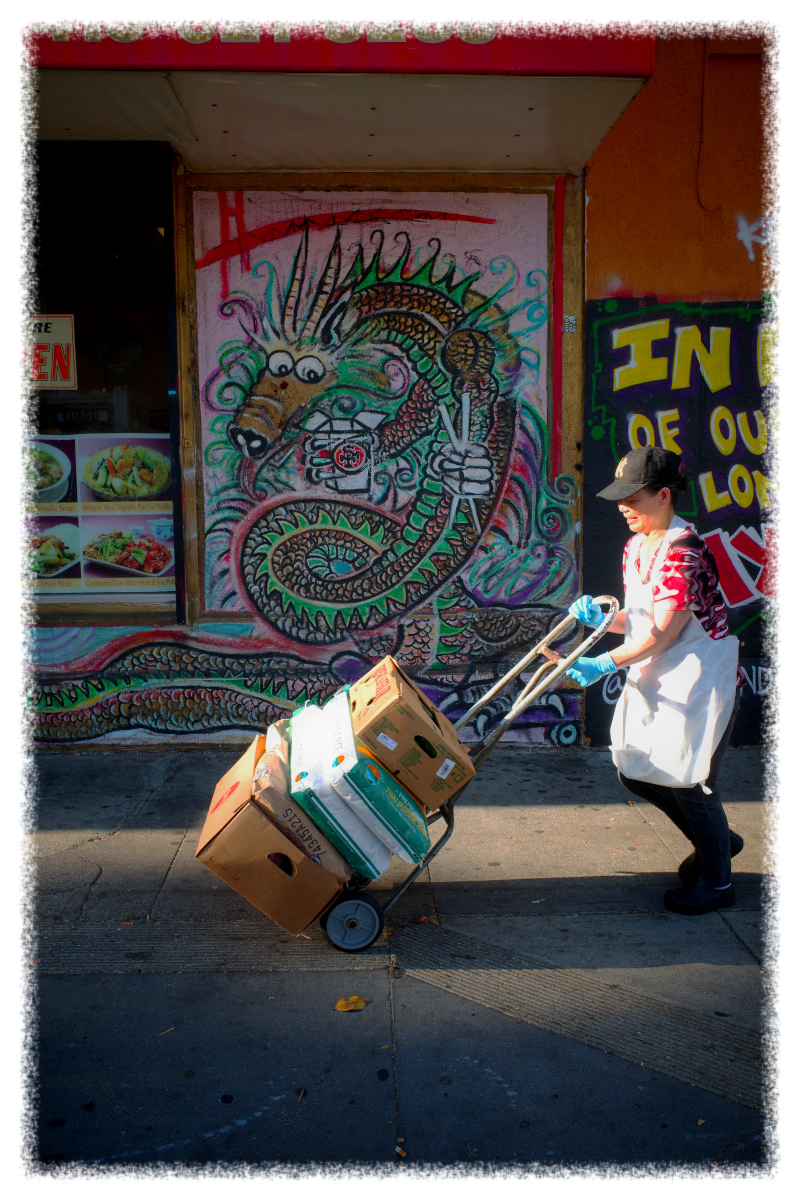 A person pushes a hand truck with boxes on a sidewalk in front of a storefront featuring a large dragon mural.