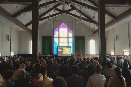 A presenter addresses an audience seated in a large room with a stained glass window projecting colorful light and a screen displaying a presentation.
