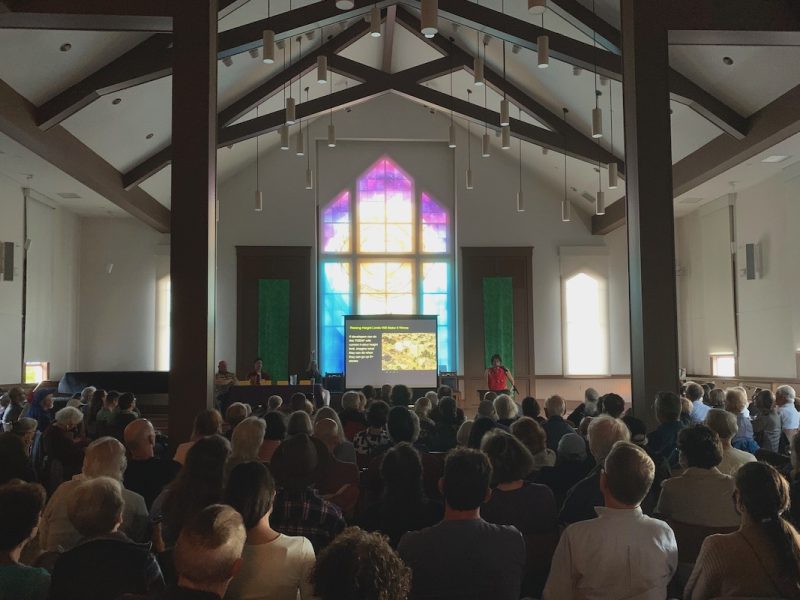 A presenter addresses an audience seated in a large room with a stained glass window projecting colorful light and a screen displaying a presentation.