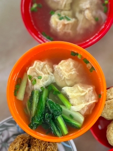 A bowl of soup with dumplings and greens in an orange bowl, accompanied by another bowl containing dumplings in broth and fried items on a plate.