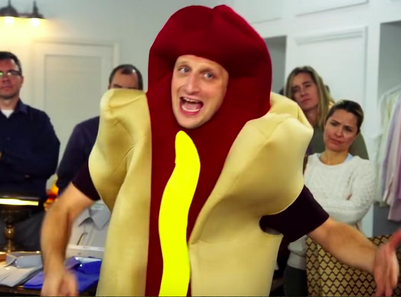 Person wearing a hot dog costume, surrounded by onlookers in a room filled with clothing racks.