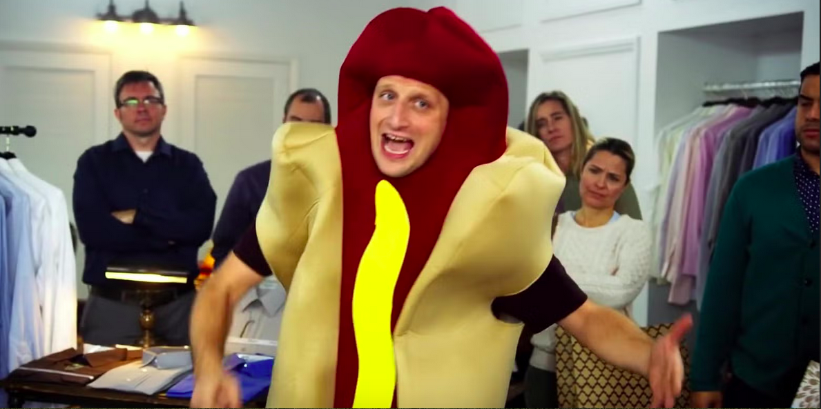 Person wearing a hot dog costume, surrounded by onlookers in a room filled with clothing racks.