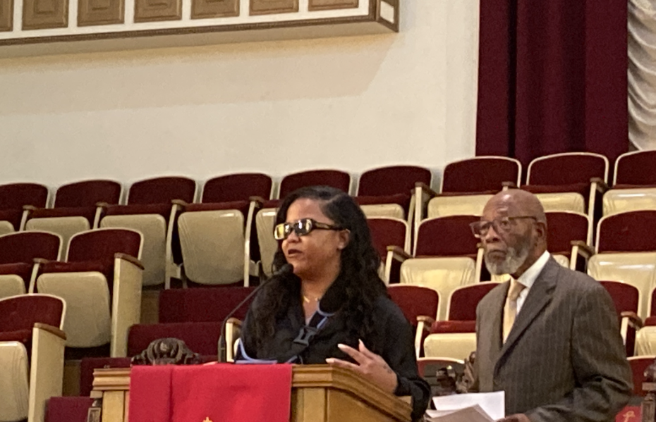 A woman speaks at a wooden podium in an auditorium with rows of empty seats, alongside an older man in a suit.