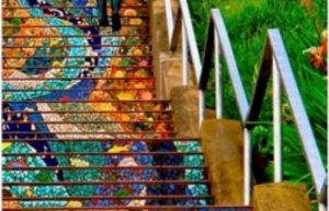 A person walks down a staircase adorned with colorful mosaic tiles, with metal railings on either side and green grass visible on the right.