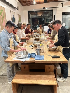 A group of people stands around a wooden table in a well-lit room, engaged in a cooking class. They are wearing aprons and handling various kitchen utensils and ingredients.